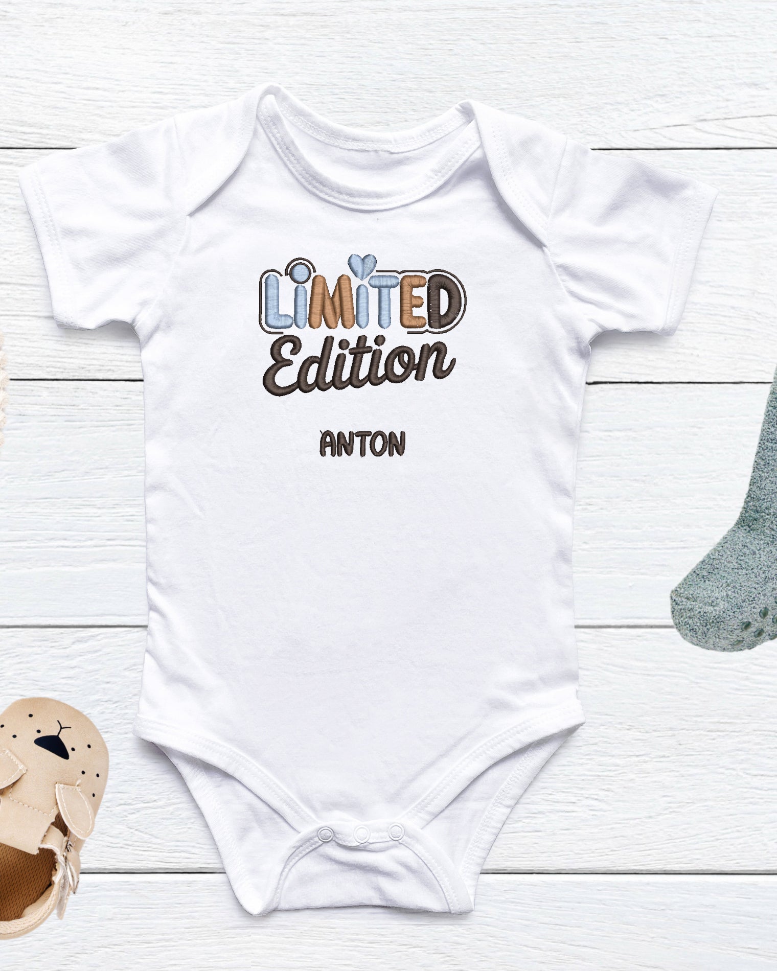 Weißer Baby-Body mit besticktem Schriftzug ‚Limited Edition‘ in Blau- und Brauntönen sowie personalisiertem Namen ‚Anton‘, auf hellem Holzuntergrund dekoriert mit Babyschuh und Stofftier