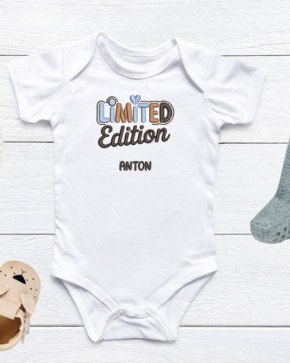 Weißer Baby-Body mit besticktem Schriftzug ‚Limited Edition‘ in Blau- und Brauntönen sowie personalisiertem Namen ‚Anton‘, auf hellem Holzuntergrund dekoriert mit Babyschuh und Stofftier