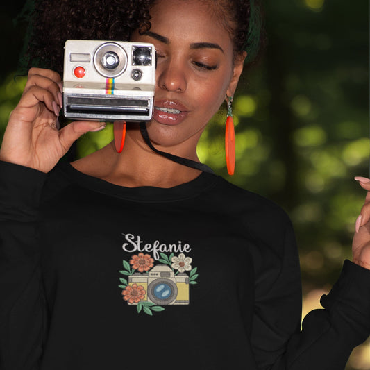 Frau hält eine Retro-Kamera und trägt ein schwarzes Sweatshirt mit besticktem Kamera- und Blumenmotiv sowie personalisiertem Namen Stefanie.