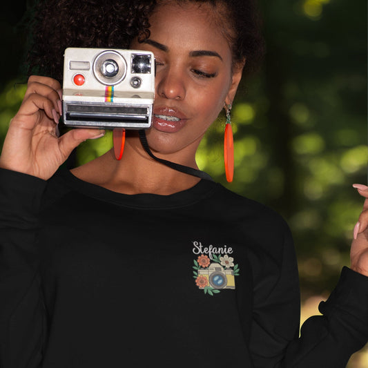 Frau hält eine Retro-Kamera und trägt ein schwarzes Sweatshirt mit besticktem Kamera- und Blumenmotiv sowie personalisiertem Namen Stefanie.